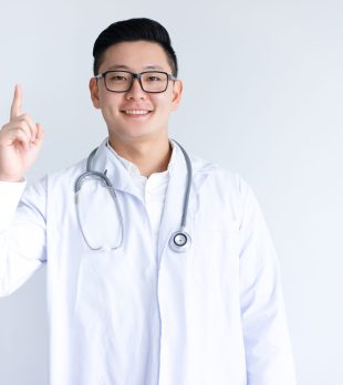 Smiling Asian male doctor pointing upwards. Man standing and looking at camera. Medical product concept. Isolated front view on white background.